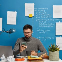 IT professional works on startup project, updates software and database on mobile phone, drinks hot beverage, sits at desktop against blue wall with written information. Computer hardware engineer