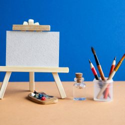 Wooden easel with textured blank paper canvas on blue brown background. Beautiful art class studio interior, watercolor brushes, pencils in a case, water. Artist's advertising poster mockup